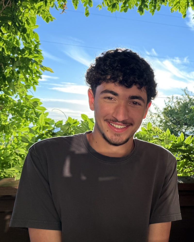 Callum, who builds AcceptMe, smiling at an outdoor table under green leaves